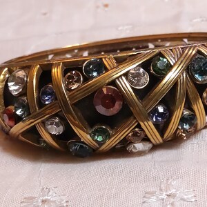 May include: A gold-toned bangle bracelet with a woven design, encrusted with a variety of colorful gemstones. The bracelet features a mix of clear, blue, green, and red stones, creating a sparkling effect. The bracelet is set against a soft, patterned background.