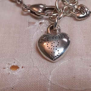 May include: A silver-toned necklace with a heart-shaped pendant. The pendant has the word "KEMA" engraved on it, along with a dotted design. The necklace is on a white, textured fabric.