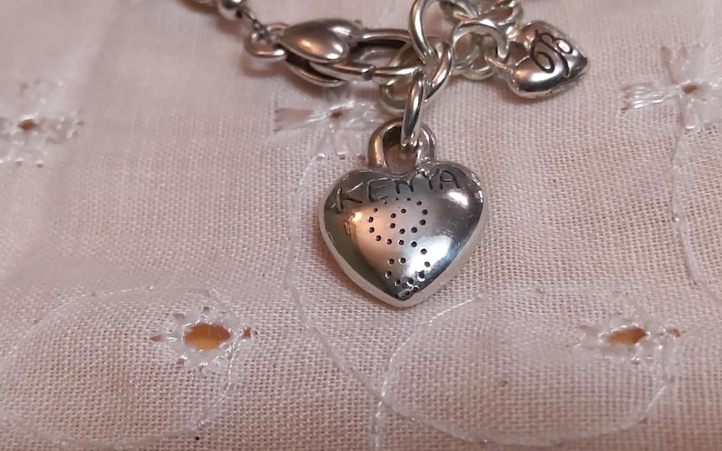 May include: A silver-toned necklace with a heart-shaped pendant. The pendant has the word "KEMA" engraved on it, along with a dotted design. The necklace is on a white, textured fabric.