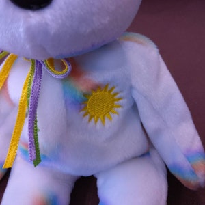 May include: White plush toy bear with a rainbow pattern and a yellow embroidered sun on the chest. The bear has a purple, yellow, and green ribbon tied around its neck.