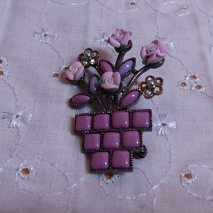 May include: A vintage brooch featuring a floral arrangement in a pot. The brooch has pink roses, green leaves, and purple accents. The pot is a square design with a dark metal frame. The background is a light pink fabric.