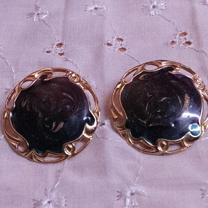 May include: A pair of vintage clip-on earrings. Each earring features a dark, swirling enamel center set within a gold-toned filigree frame. The earrings are round and have a classic, elegant design.