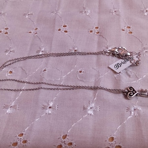 Op de afbeelding: Zilverkleurige ketting met een sleutelhanger. De sleutel heeft een hartvormige bovenkant en een gedetailleerd ontwerp. De ketting ligt op een witte, bedrukte stof. Een merklabel is bevestigd.