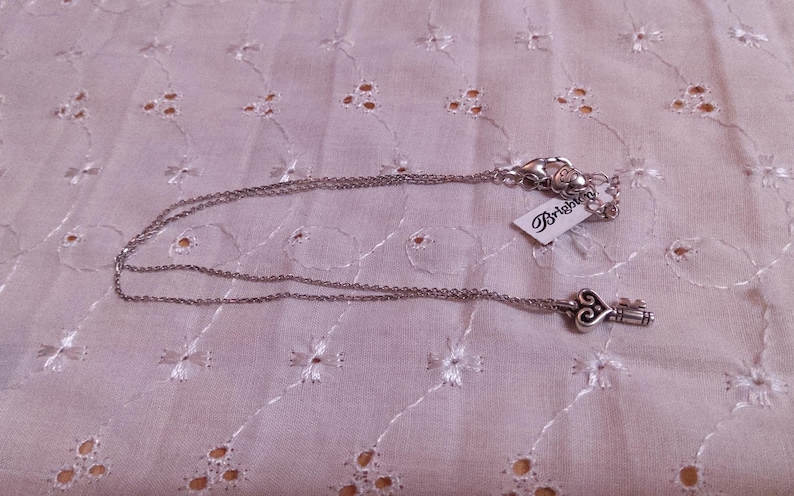 Op de afbeelding: Zilverkleurige ketting met een sleutelhanger. De sleutel heeft een hartvormige bovenkant en een gedetailleerd ontwerp. De ketting ligt op een witte, bedrukte stof. Een merklabel is bevestigd.