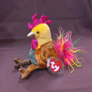 May include: A plush rooster toy with a yellow body, brown and green wings, and a pink and green feathered tail. The rooster has a pink comb and wattle, and a silver beak. The toy has a Ty tag attached to it.