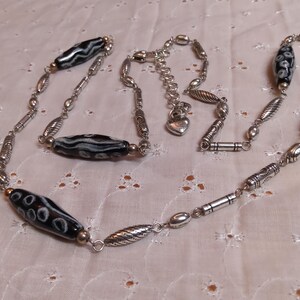 Op de afbeelding: Een zilverkleurige ketting met langwerpige zwart-wit patronen kralen. De ketting heeft diverse zilverkleurige metalen kralen en een hartvormige bedel. De ketting wordt gepresenteerd op een witte, getextureerde ondergrond.