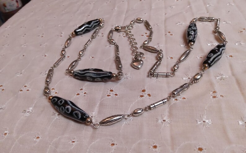 Op de afbeelding: Een zilverkleurige ketting met langwerpige zwart-wit patronen kralen. De ketting heeft diverse zilverkleurige metalen kralen en een hartvormige bedel. De ketting wordt gepresenteerd op een witte, getextureerde ondergrond.