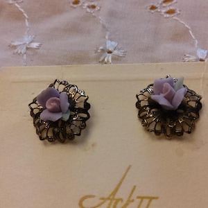 May include: A pair of vintage earrings featuring a delicate floral design. Each earring has a lavender-colored rose at the center, surrounded by a filigree metal frame. The earrings are displayed on a cream-colored card.