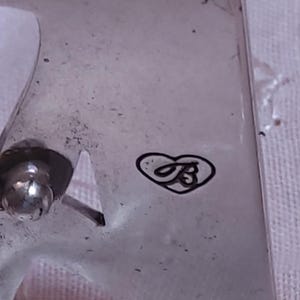 May include: Close-up of a silver-toned metal object with a heart-shaped logo containing the letter "B". The object has a rounded, ball-shaped element and a cut-out design. The surface appears slightly textured.