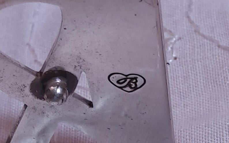 May include: Close-up of a silver-toned metal object with a heart-shaped logo containing the letter "B". The object has a rounded, ball-shaped element and a cut-out design. The surface appears slightly textured.