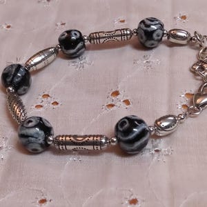 May include: A beaded bracelet featuring black and white patterned beads, silver-toned metal beads, and a heart charm. The bracelet is approximately 7 inches long and is displayed on a white, floral-patterned fabric.