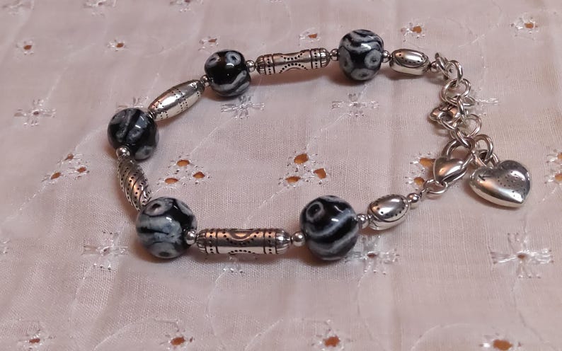 May include: A beaded bracelet featuring black and white patterned beads, silver-toned metal beads, and a heart charm. The bracelet is approximately 7 inches long and is displayed on a white, floral-patterned fabric.