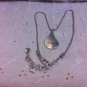 May include: A silver necklace with a teardrop-shaped pendant featuring a swirling design. The necklace has a lobster clasp and a small heart charm.