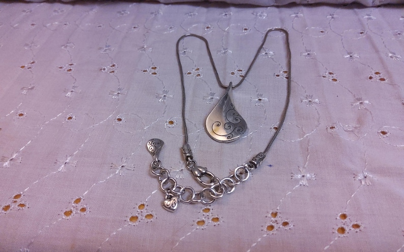 May include: A silver necklace with a teardrop-shaped pendant featuring a swirling design. The necklace has a lobster clasp and a small heart charm.