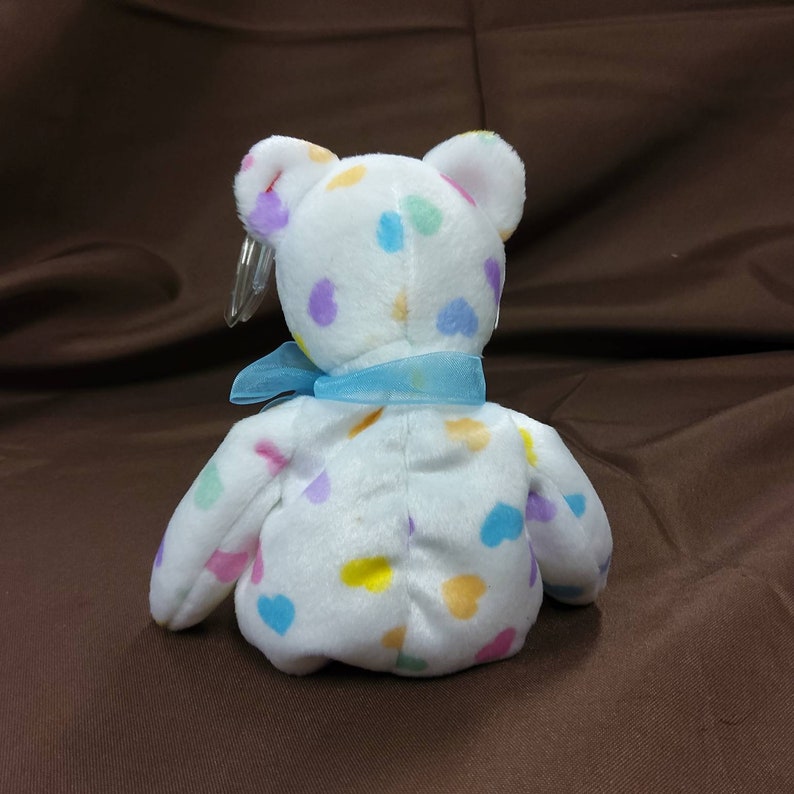 May include: White plush teddy bear with a blue ribbon around its neck. The bear is covered in colorful heart-shaped spots.