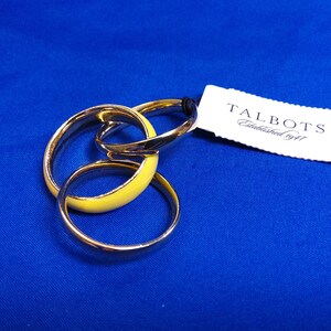 May include: A set of three interlocking gold-tone rings, one with a yellow enamel finish, on a blue background. A white tag with the brand name "TALBOTS" and "Established 1947" is attached to the rings.