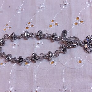 May include: A silver bracelet with a heart-shaped charm. The charm is decorated with clear rhinestones. The bracelet is made of a chain with small silver beads.