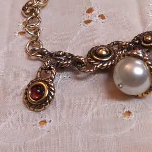 May include: A vintage-style gold-toned bracelet with ornate detailing. The bracelet features a large, lustrous pearl and a small, oval-shaped red gemstone pendant. The chain links are textured, and the clasp is visible.