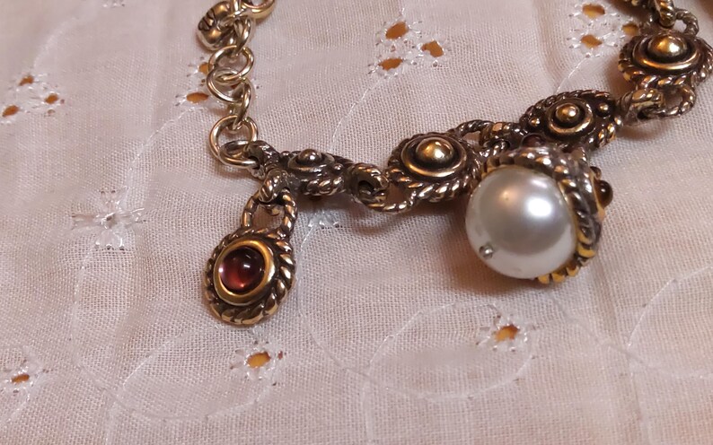 May include: A vintage-style gold-toned bracelet with ornate detailing. The bracelet features a large, lustrous pearl and a small, oval-shaped red gemstone pendant. The chain links are textured, and the clasp is visible.