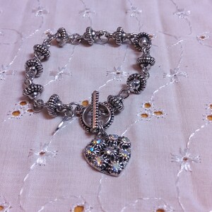 May include: A silver charm bracelet with a heart-shaped charm. The heart charm is decorated with small, clear rhinestones. The bracelet is made of a chain with small, round beads.