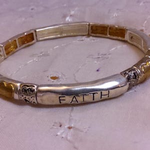 May include: Silver bracelet with gold accents and the word "FAITH" engraved on the front. The bracelet is made of small beads and has a stretchy design.