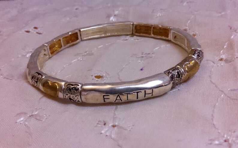 May include: Silver bracelet with gold accents and the word "FAITH" engraved on the front. The bracelet is made of small beads and has a stretchy design.