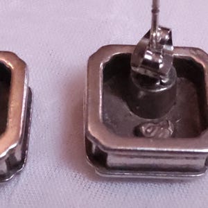 May include: Close-up of two silver-toned square stud earrings. The earrings have a raised, rectangular border and a post with a butterfly clasp. The earrings are on a white surface.