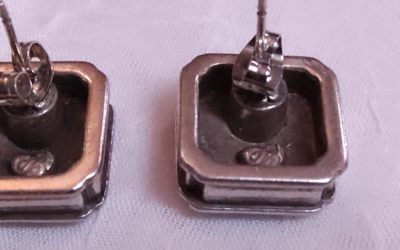 May include: Close-up of two silver-toned square stud earrings. The earrings have a raised, rectangular border and a post with a butterfly clasp. The earrings are on a white surface.