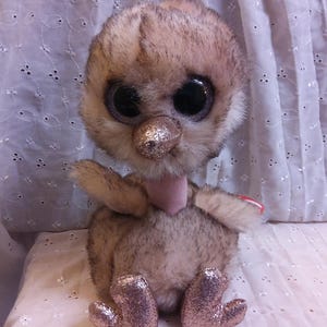 May include: A plush toy ostrich with large, glittery eyes, a pink beak, and pink glitter on its feet. The toy is sitting on a white fabric background.