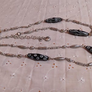Op de afbeelding: Een zilverkleurige ketting met langwerpige zwarte kralen met witte cirkelvormige patronen. De ketting heeft een reeks kleinere zilveren kralen en decoratieve elementen, met een verstelbare ketting en een hartvormige bedel.