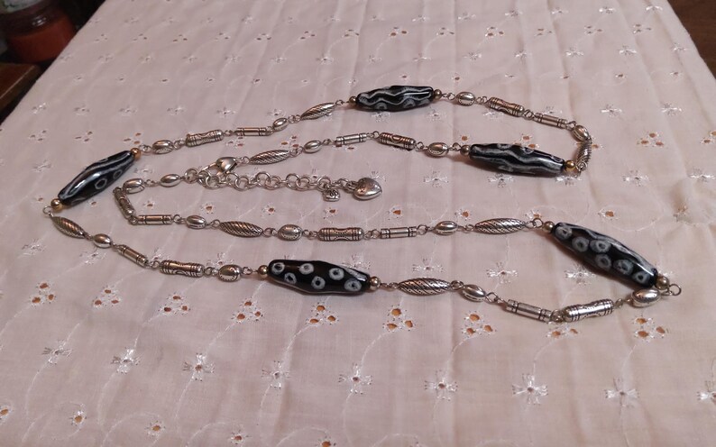 Op de afbeelding: Een zilverkleurige ketting met langwerpige zwarte kralen met witte cirkelvormige patronen. De ketting heeft een reeks kleinere zilveren kralen en decoratieve elementen, met een verstelbare ketting en een hartvormige bedel.