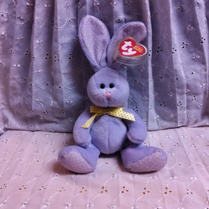May include: Purple plush bunny with yellow polka dots on its ears and feet. It has a yellow ribbon around its neck and a red Ty Beanie Baby tag attached to its ear. The bunny is sitting on a light purple fabric background.