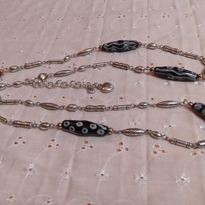 Op de afbeelding: Lange zilveren ketting met afwisselend zwart-witte kralen met patronen en zilverkleurige metalen accenten. De ketting heeft ovale en cilindrische kralen, met een hartvormige sluiting.