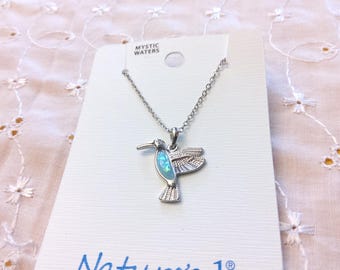 May include: Silver-tone hummingbird pendant necklace with a blue-green opal-like center. The necklace hangs on a delicate chain and is displayed on a white card with the text "Mystic Waters" and "Nature's 1".