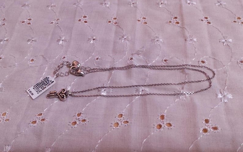 Op de afbeelding: Zilverkleurige ketting met een sleutelhanger. De sleutelhanger heeft een hartvormig ontwerp. De ketting ligt op een witte stof met een bloemenpatroon. Een prijskaartje is bevestigd.