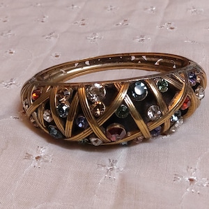 May include: A gold-toned bangle bracelet with a woven design. The bracelet is adorned with a variety of colorful gemstones, including clear, red, blue, and green. The bracelet is a vintage piece.