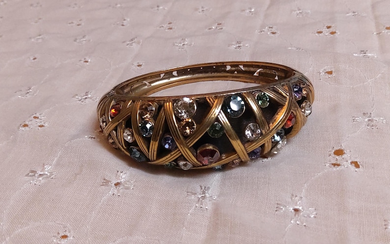 May include: A gold-toned bangle bracelet with a woven design. The bracelet is adorned with a variety of colorful gemstones, including clear, red, blue, and green. The bracelet is a vintage piece.