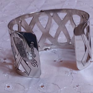 May include: Silver-toned cuff bracelet with an openwork design featuring intersecting ribbon-like shapes. The bracelet has a curved shape and the word "Brighton" is visible on the side.