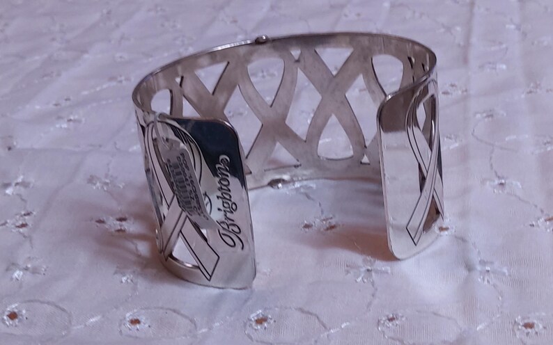 May include: Silver-toned cuff bracelet with an openwork design featuring intersecting ribbon-like shapes. The bracelet has a curved shape and the word "Brighton" is visible on the side.