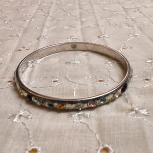 May include: Silver-toned bangle bracelet with a row of colorful gemstones. The bracelet has a simple design with a smooth, polished finish. The gemstones are various colors, including blue, pink, purple, and yellow. A small heart-shaped design is visible on the bracelet.