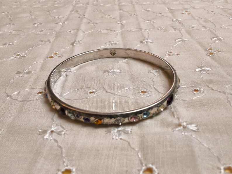 May include: Silver-toned bangle bracelet with a row of colorful gemstones. The bracelet has a simple design with a smooth, polished finish. The gemstones are various colors, including blue, pink, purple, and yellow. A small heart-shaped design is visible on the bracelet.