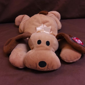 May include: A brown plush dog toy with a white bow and a red Ty tag. The dog is lying on its back with its legs in the air.