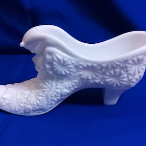 May include: A white, decorative shoe-shaped dish. The dish is made of glass or ceramic and features a detailed, embossed floral pattern. The shoe has a small heel and a curved opening, perfect for display or small items.