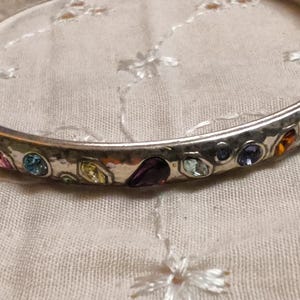 May include: Silver-toned bangle bracelet with a hammered texture, adorned with colorful gemstones. The gemstones include pink, blue, yellow, purple, and orange hues. The bracelet is set against a white fabric background.