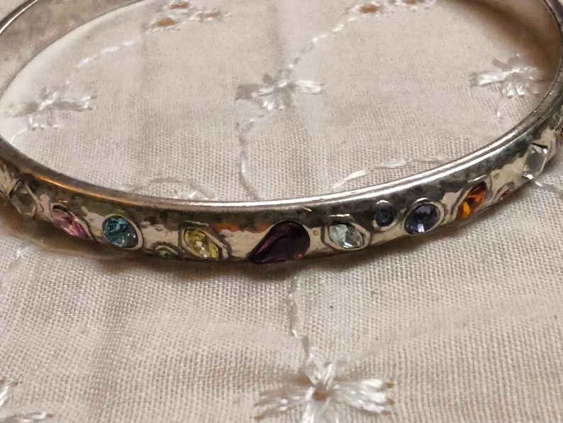 May include: Silver-toned bangle bracelet with a hammered texture, adorned with colorful gemstones. The gemstones include pink, blue, yellow, purple, and orange hues. The bracelet is set against a white fabric background.