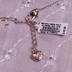 Op de afbeelding: Zilverkleurige ketting met een hartvormige bedel en een label met de tekst "MICRO MNI KEY JL2892 SZ: OS". De ketting ligt op een roze, geborduurde stof.