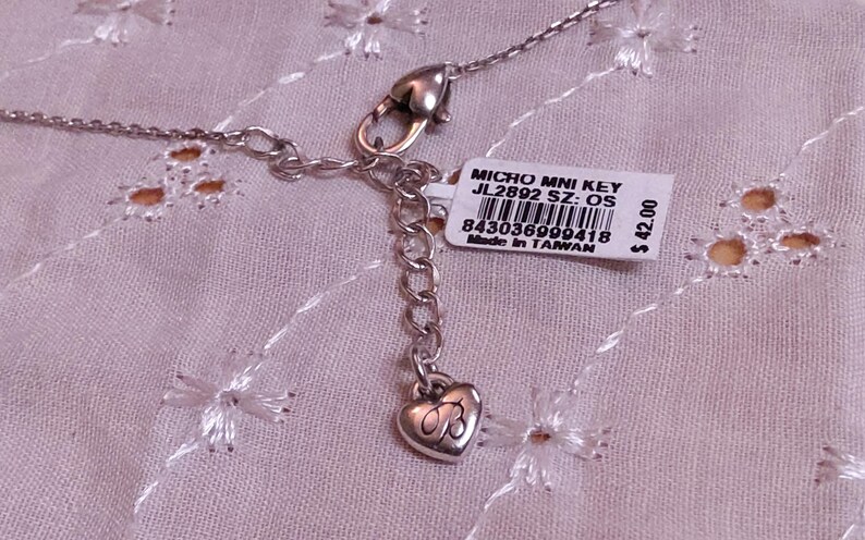 Op de afbeelding: Zilverkleurige ketting met een hartvormige bedel en een label met de tekst "MICRO MNI KEY JL2892 SZ: OS". De ketting ligt op een roze, geborduurde stof.