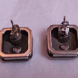 May include: Two silver-toned square stud earrings with a gold-colored border. The backs of the earrings are visible, showing the post and butterfly clasp. The earrings are on a light purple surface.