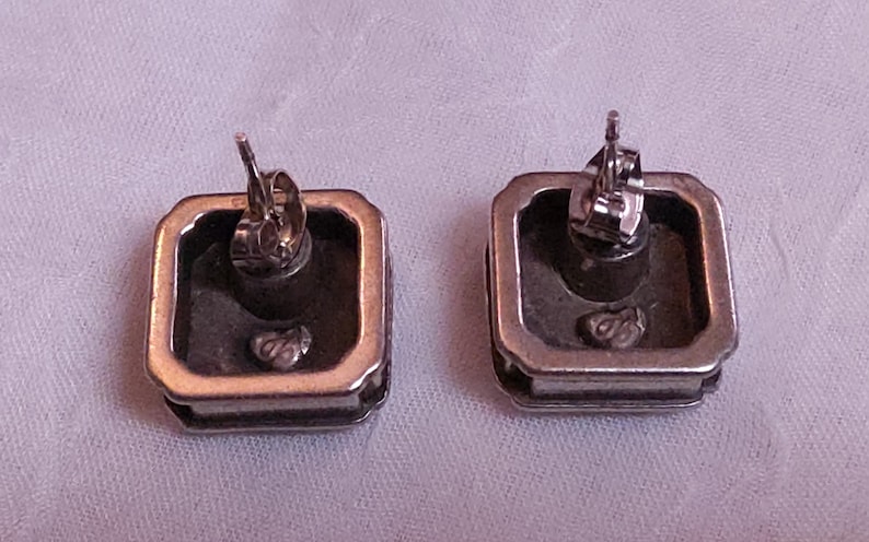 May include: Two silver-toned square stud earrings with a gold-colored border. The backs of the earrings are visible, showing the post and butterfly clasp. The earrings are on a light purple surface.