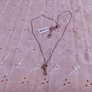 Op de afbeelding: Een zilverkleurige ketting met een sleutelhanger. De sleutelhanger heeft een hartvormige bovenkant en een klein sleutelontwerp. De ketting heeft een label met de tekst "Brighton". De ketting wordt getoond op een lichtroze, geborduurde stof.